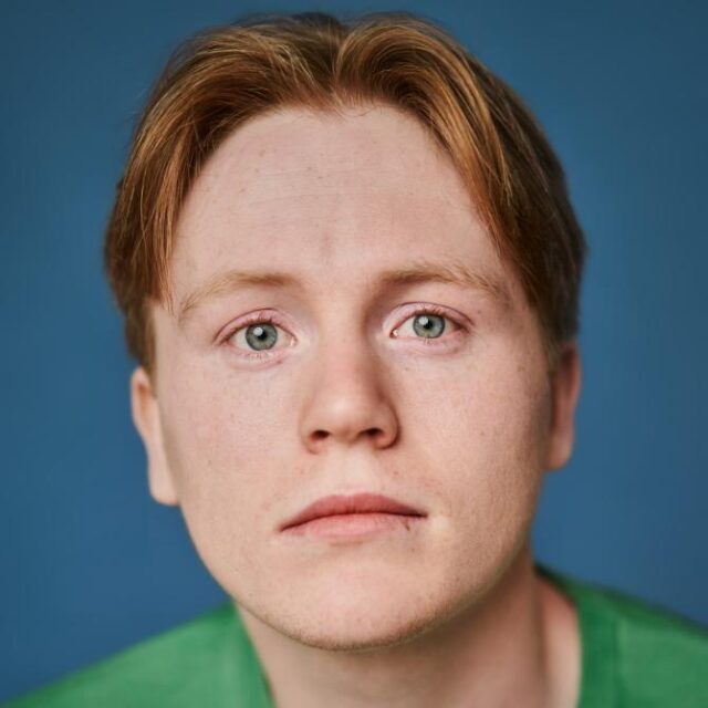 Close-up portrait of a young man with red hair and green eyes, wearing a green top, against a blue background