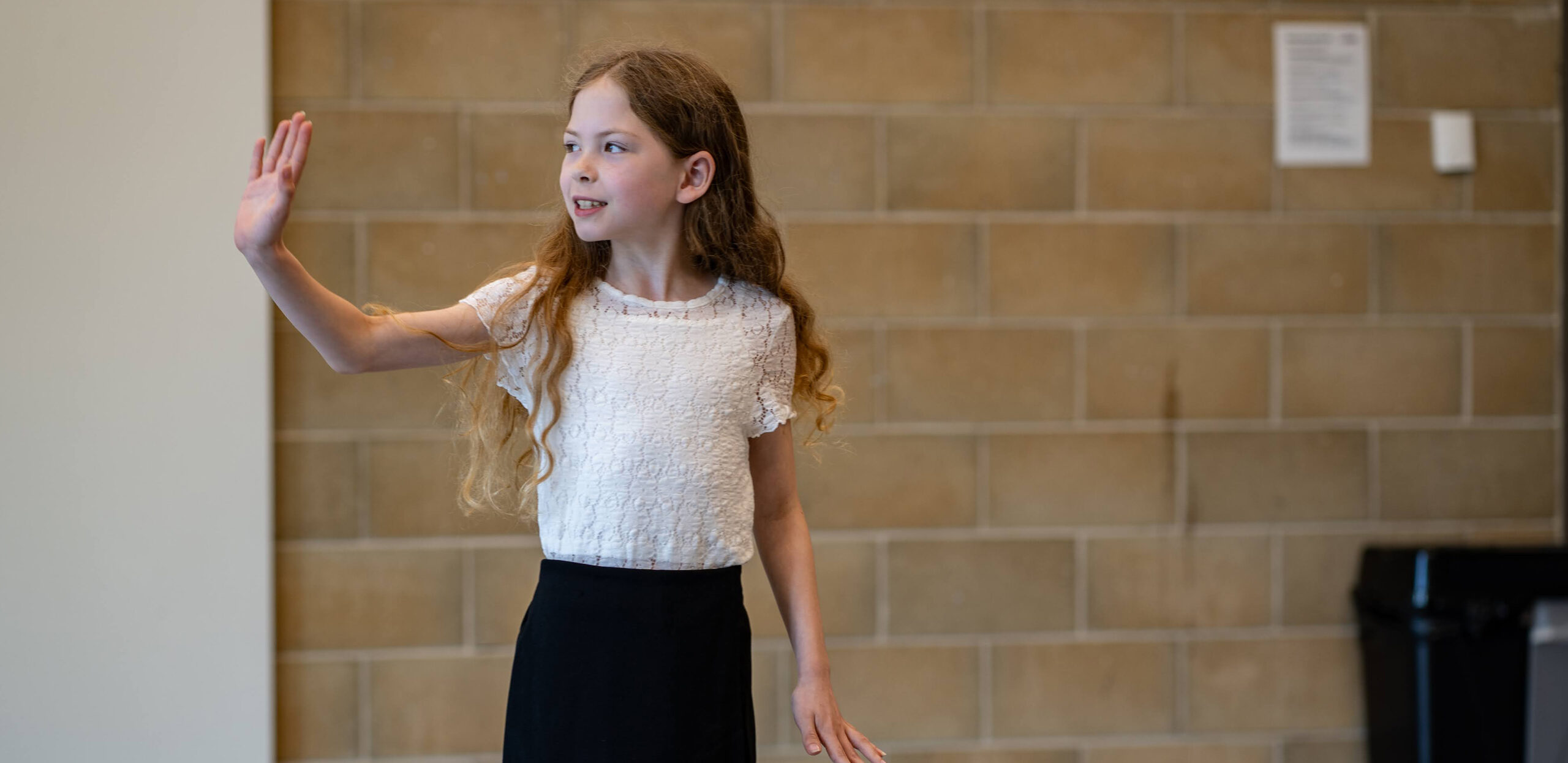 Young girl with long hair raises her hand and speaks while standing in a corridor