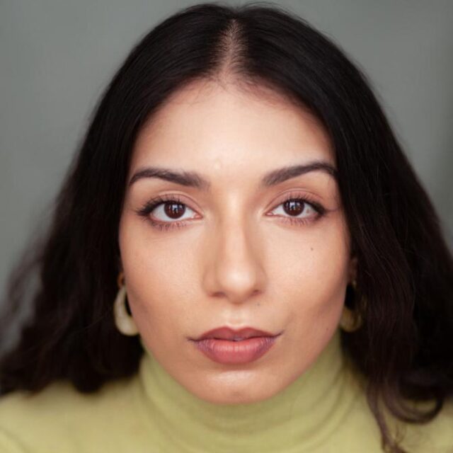 Close-up portrait of a woman with dark wavy hair, hoop earrings and a pale green polo neck jumper