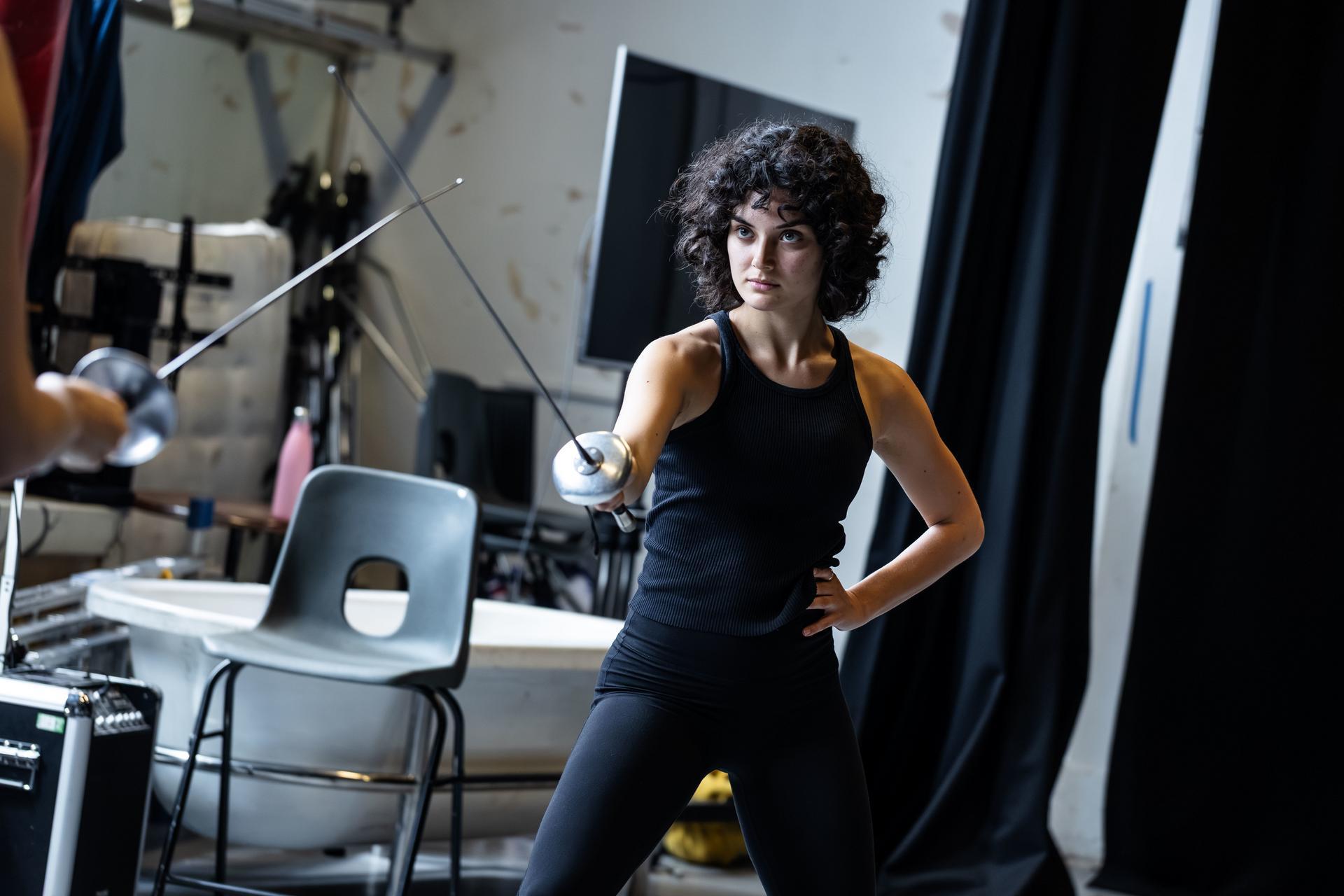 Woman with curly hair holding a fencing sword in a lunge stance, crossing blades with an opponent's foil