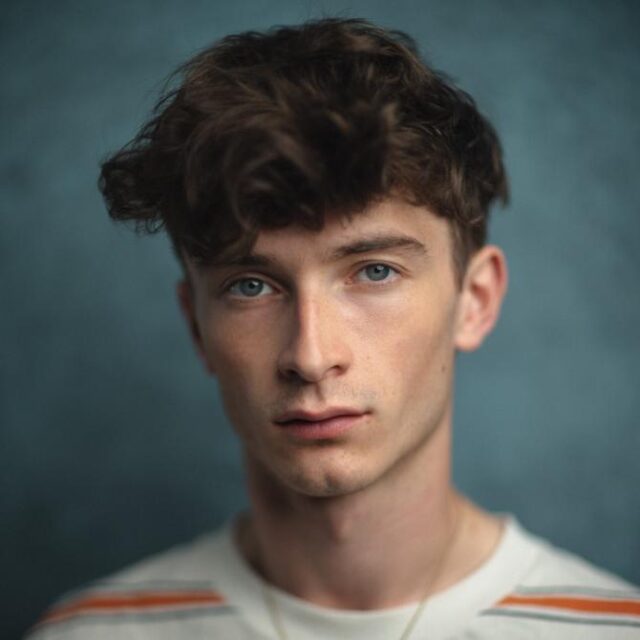 Portrait of a young man with curly brown hair and blue eyes, looking directly at the camera against a teal backdrop
