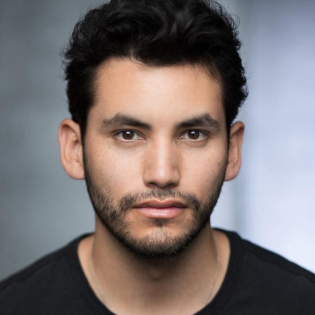 Headshot of a man with curly dark hair and stubble, wearing a black top, against a grey background