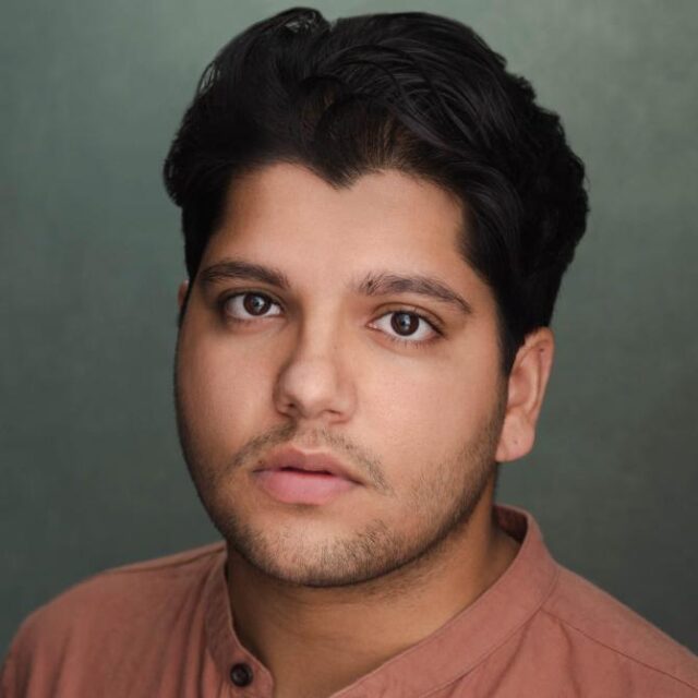 Studio portrait of a young man with dark hair and light stubble, wearing a terracotta shirt, against a green backdrop