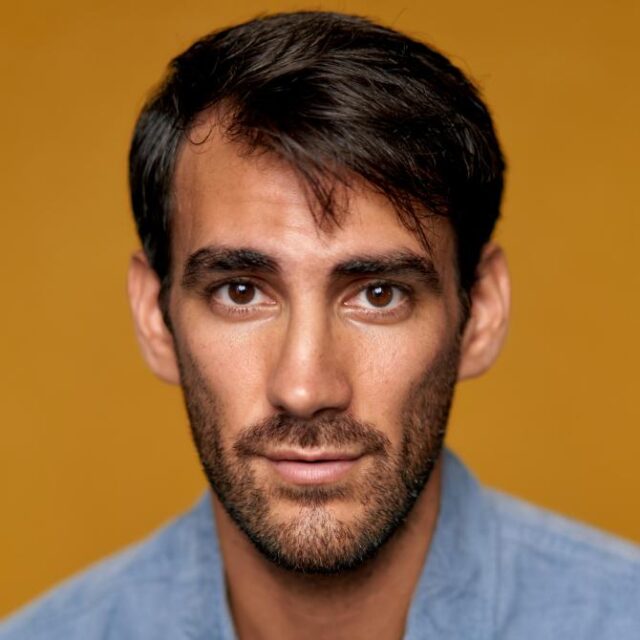 Close-up portrait of a man with dark hair and a short beard, wearing a light blue shirt, against a mustard background