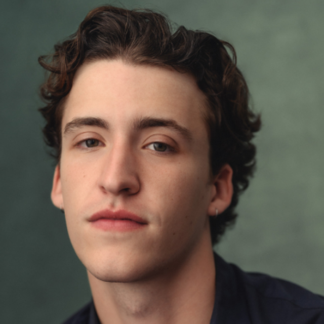 Portrait of a young man with curly brown hair and a small stud earring, looking directly at the camera