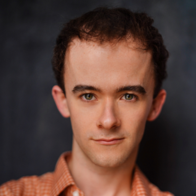 Headshot of a young man with curly brown hair and green eyes, wearing an orange checked shirt, against a dark background
