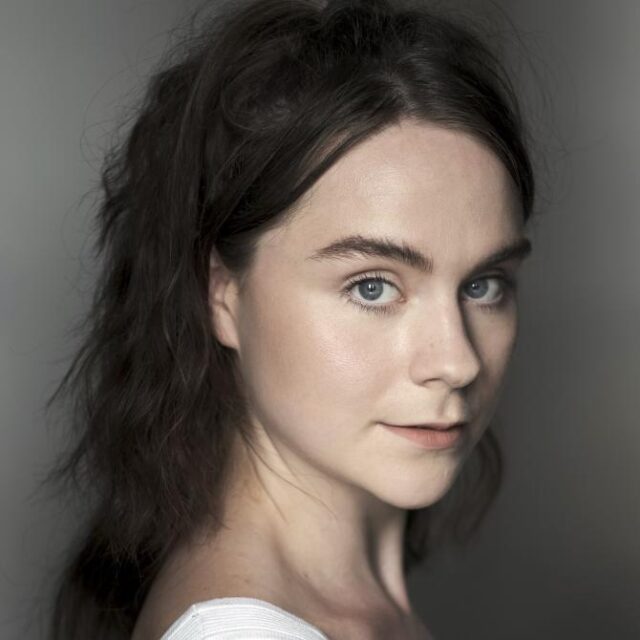 Close-up portrait of a young woman with wavy dark hair, looking over her shoulder at the camera