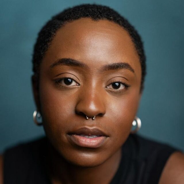 Close-up portrait of a woman with short natural hair, septum piercing and hoop earrings, looking directly at the camera
