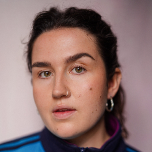 Close-up portrait of a young woman with dark hair tied back, a nose ring and hoop earring, wearing a blue tracksuit top