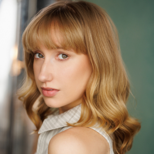 Woman with wavy blonde hair and fringe, looking over her shoulder at the camera