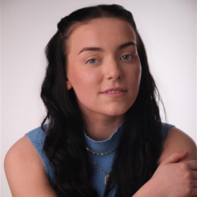 Young woman with long dark hair, wearing a blue top and layered necklaces, looking at the camera