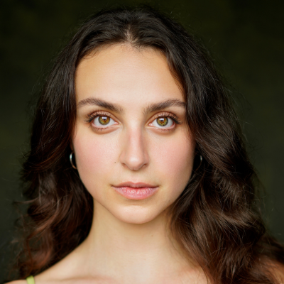 Close-up portrait of a young woman with long wavy dark hair, looking directly at the camera against a dark green background