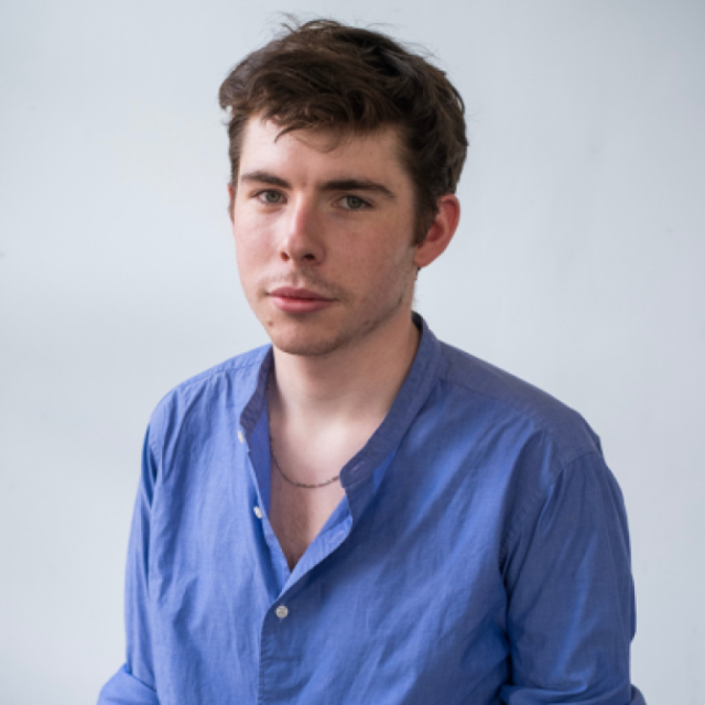 Portrait of a young man with wavy brown hair, wearing a blue collarless shirt, against a plain grey background