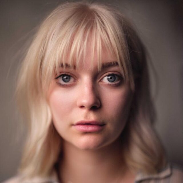 Close-up portrait of a young woman with blonde fringe and grey-blue eyes, looking directly at the camera