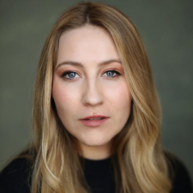 Close-up portrait of a woman with long blonde hair and blue eyes, wearing subtle makeup, against a plain grey-green background