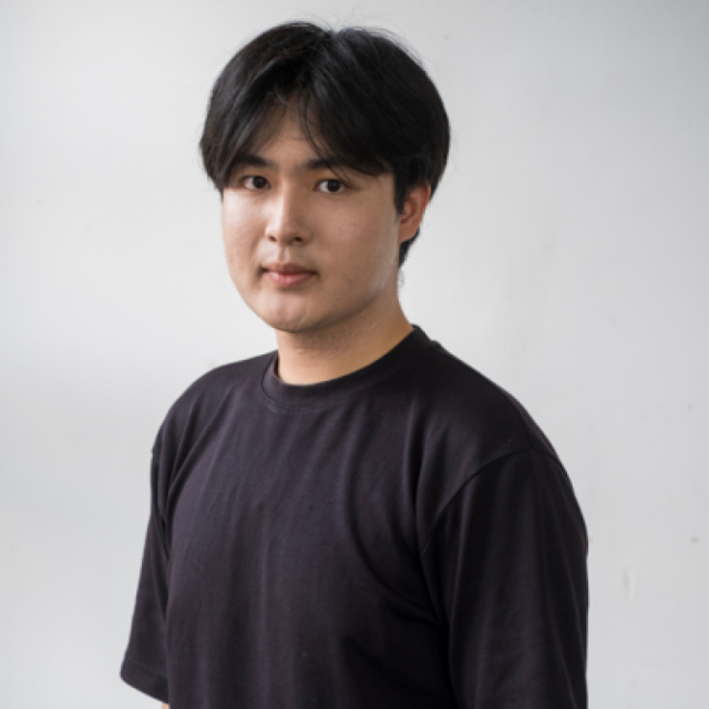 Portrait of a young man with dark hair wearing a black T-shirt against a plain grey background