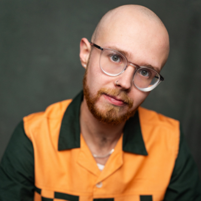 Bald man with a ginger beard and round glasses, wearing an orange and dark green shirt