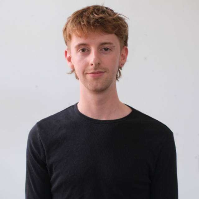 Portrait of a young man with ginger hair and earrings, wearing a black jumper, against a plain grey background