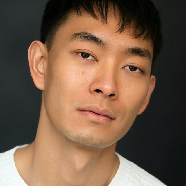Close-up portrait of a young man with short dark hair, wearing a white top, against a dark background