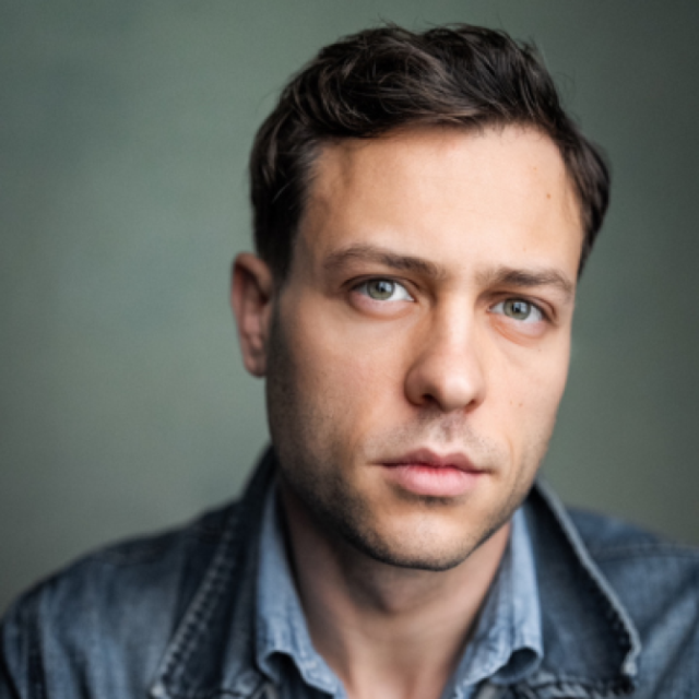 Portrait of a man with dark wavy hair wearing a denim jacket, looking directly at the camera