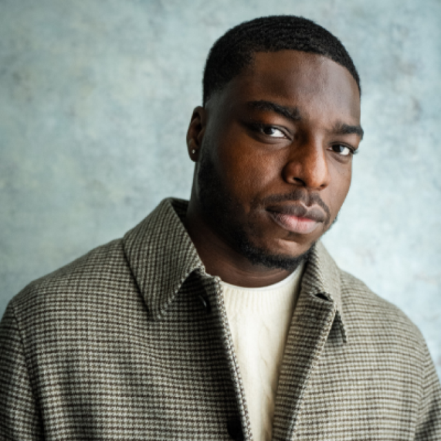Man with short hair and beard, wearing a checked coat over a cream jumper, looking at the camera
