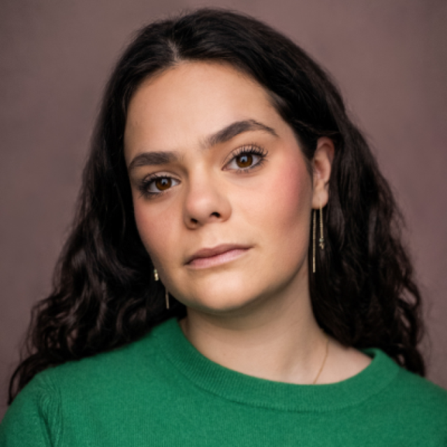 Woman with long curly dark hair wearing a green jumper and dangling earrings, looking at the camera