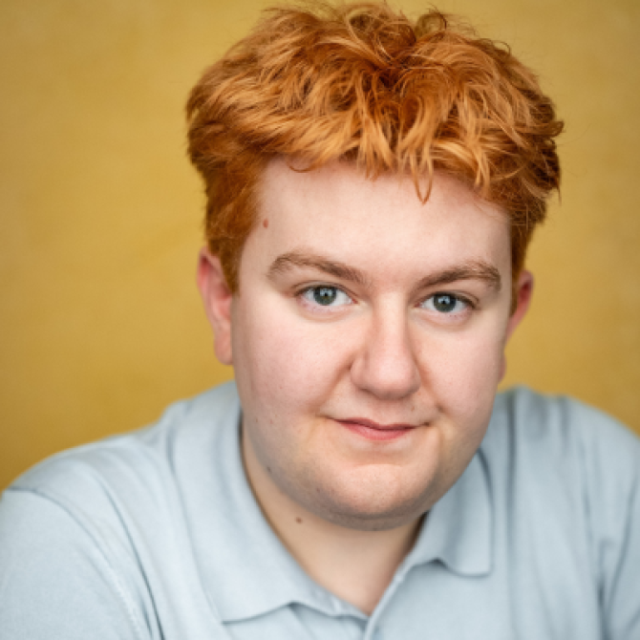 Man with curly red hair wearing a light grey polo shirt, smiling against a mustard-yellow background