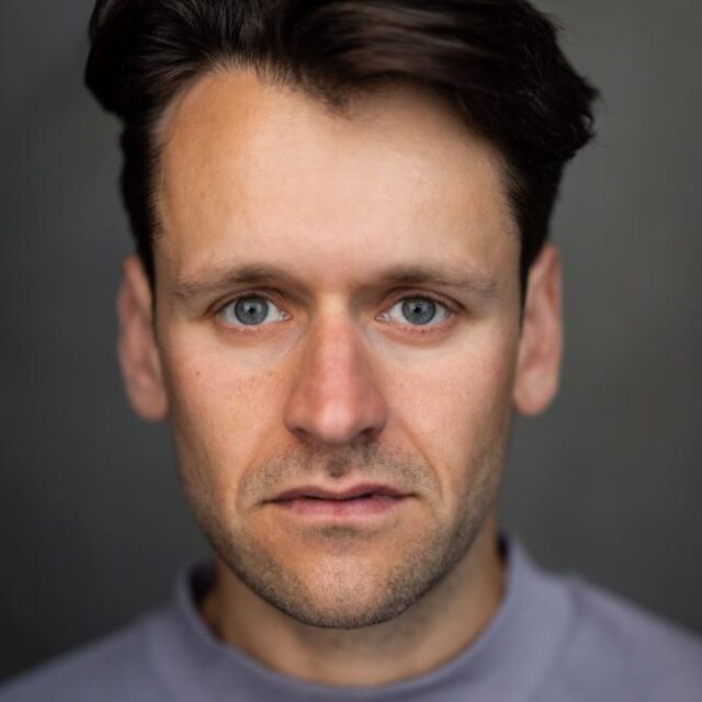 Close-up portrait of a man with dark hair and blue eyes, looking directly at the camera with a neutral expression