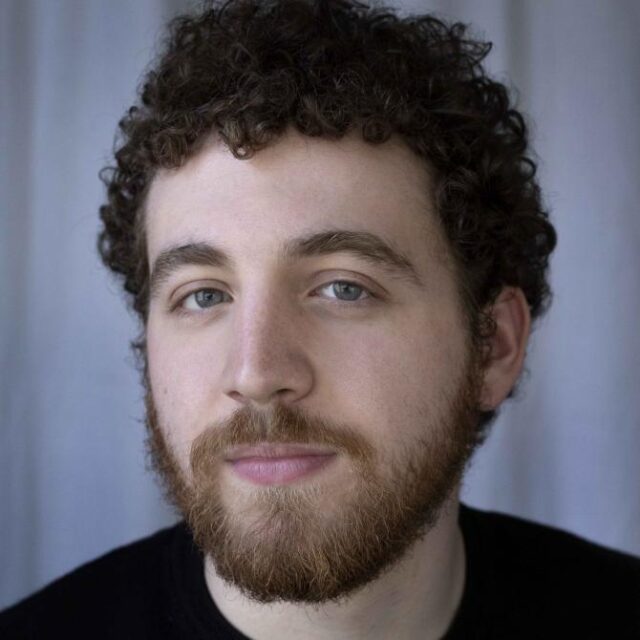 Man with curly hair and a beard looking directly at the camera against a light backdrop