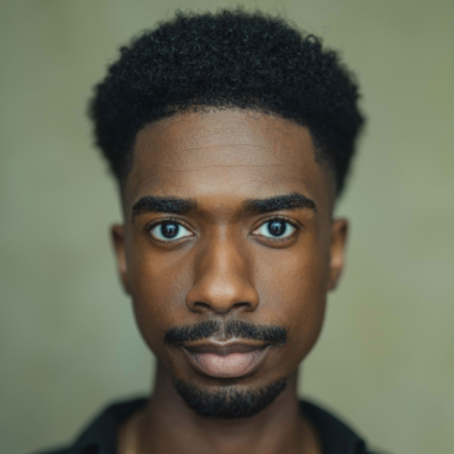 Close-up portrait of a young man with a moustache and beard, looking directly at the camera