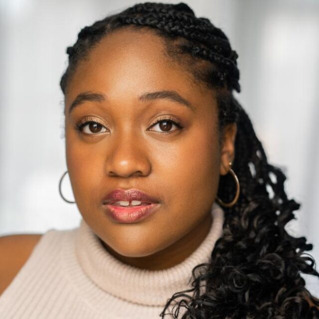 Close-up portrait of a woman with braided hair and hoop earrings, wearing a cream turtleneck, looking directly at the camera