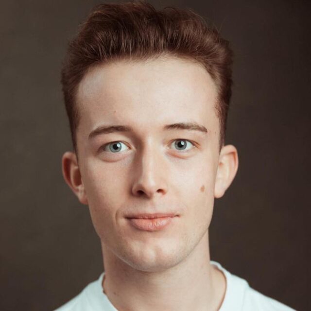 Headshot of a young man with reddish-brown hair and blue eyes, wearing a white top, against a brown background