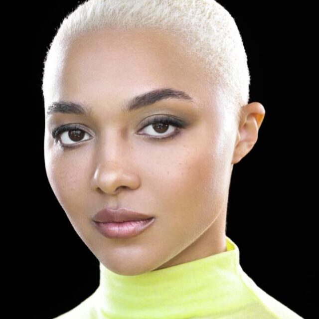 Close-up portrait of a woman with short bleached blonde hair, defined eye make-up and nude lipstick, wearing a neon yellow polo neck