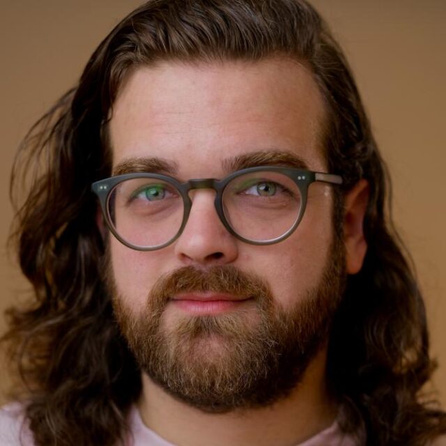 Man with long wavy brown hair, round glasses and a full beard, smiling gently