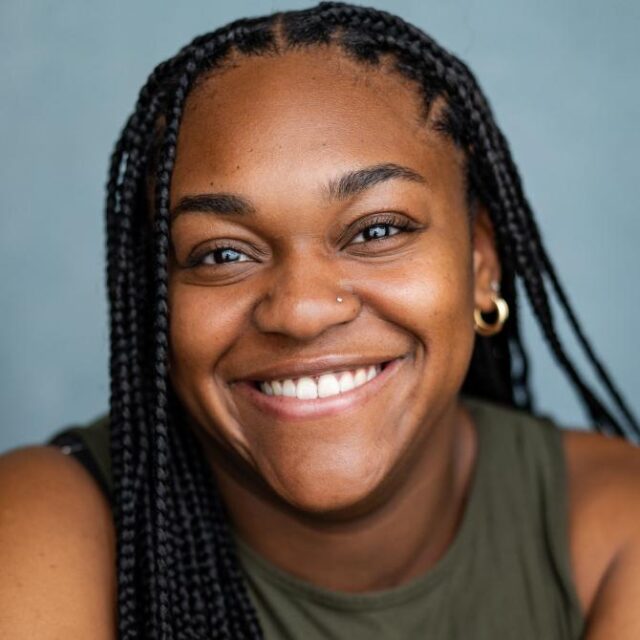 Close-up portrait of a smiling woman with long braided hair, a nose stud and hoop earring, wearing an olive green top