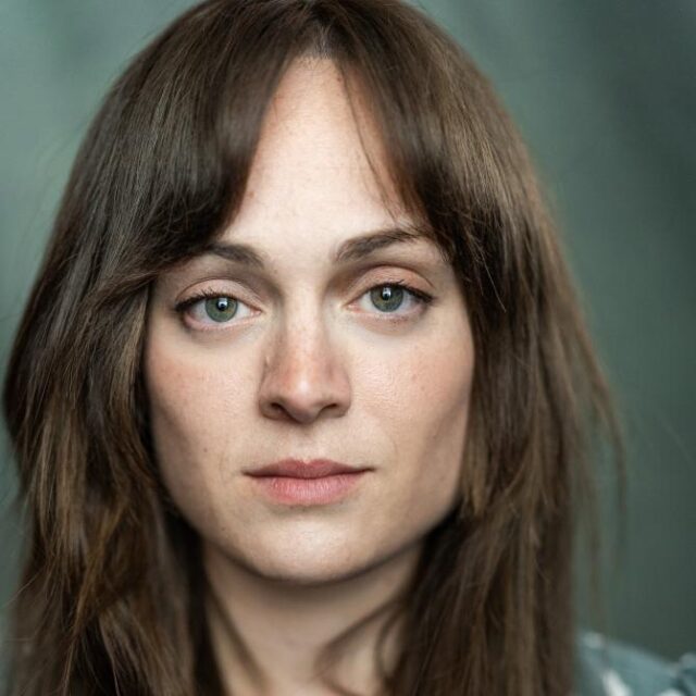 Close-up portrait of a woman with long brown hair and fringe, looking directly at the camera with a neutral expression