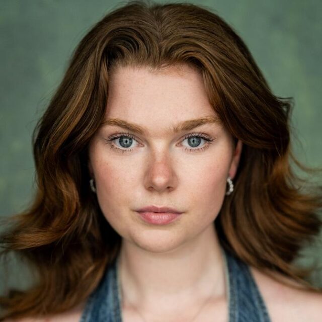 Close-up portrait of a young woman with wavy auburn hair and blue-green eyes, looking directly at the camera
