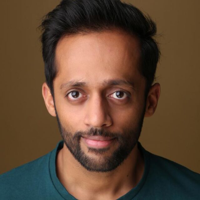 Headshot of a man with dark hair and a beard, wearing a teal top, smiling slightly at the camera