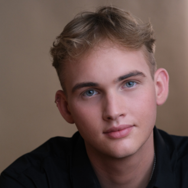 Close-up portrait of a young man with blond hair and blue eyes, wearing a dark shirt