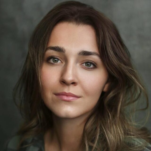 Close-up portrait of a young woman with wavy brown hair, looking directly at the camera against a grey background