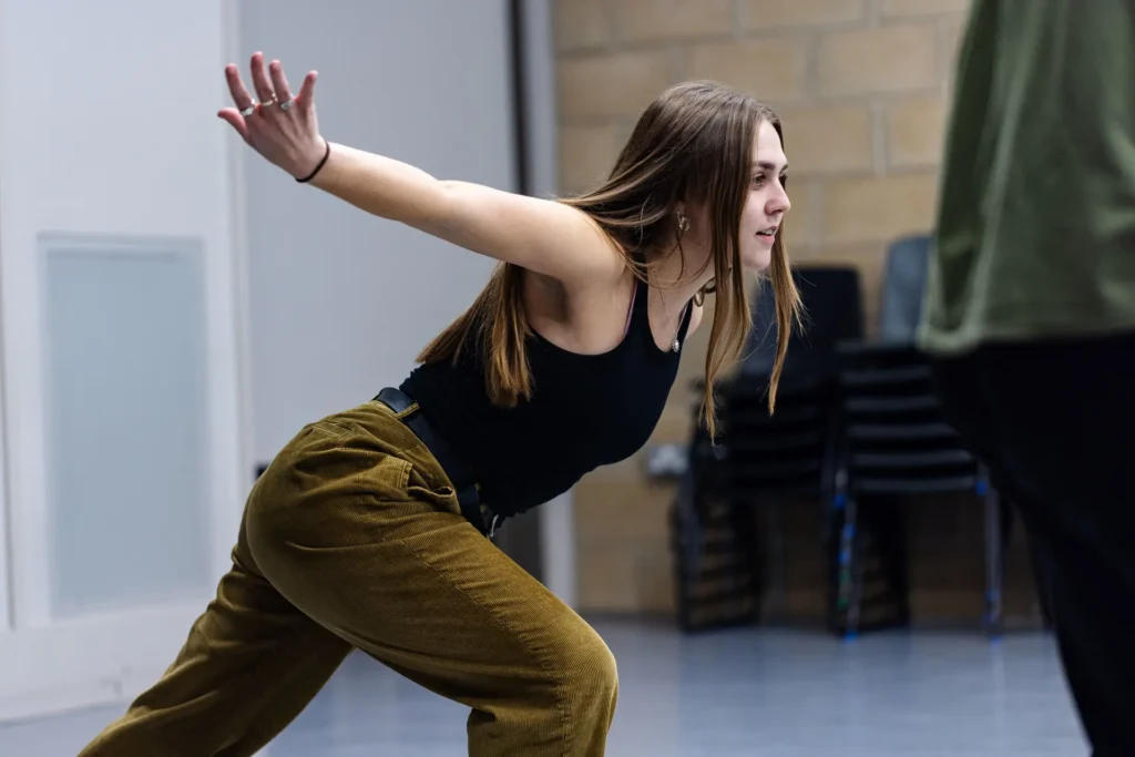 A woman in a black top and khaki trousers dances mid-lunge, arm extended, during a rehearsal in a studio
