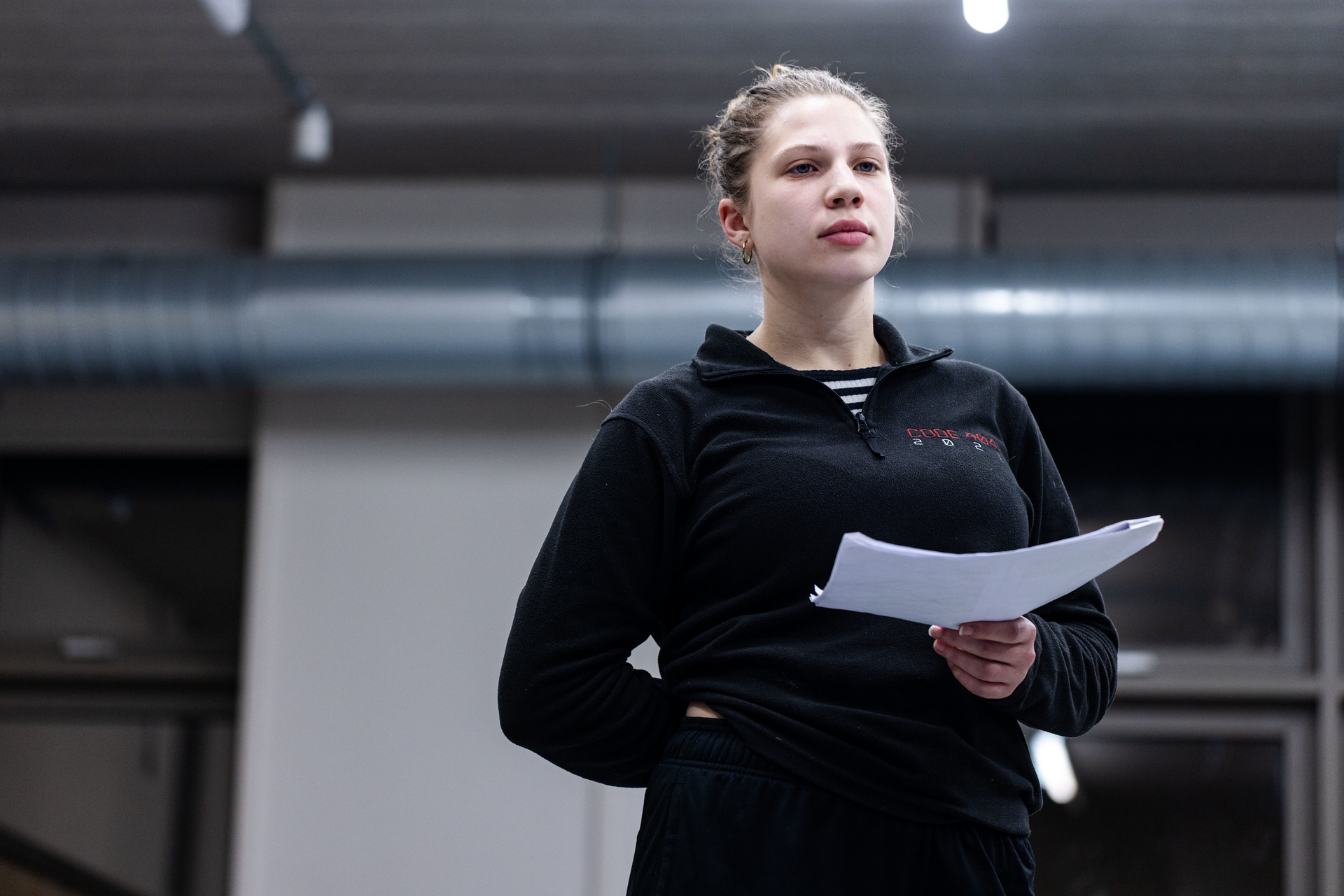Young woman in a black fleece holding papers, standing indoors in front of exposed ductwork