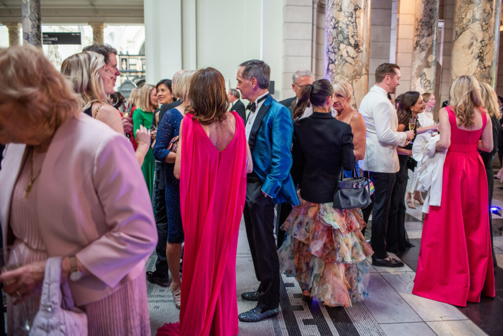 Guests in formal evening wear mingle at a reception in a marble-columned gallery, holding glasses of champagne