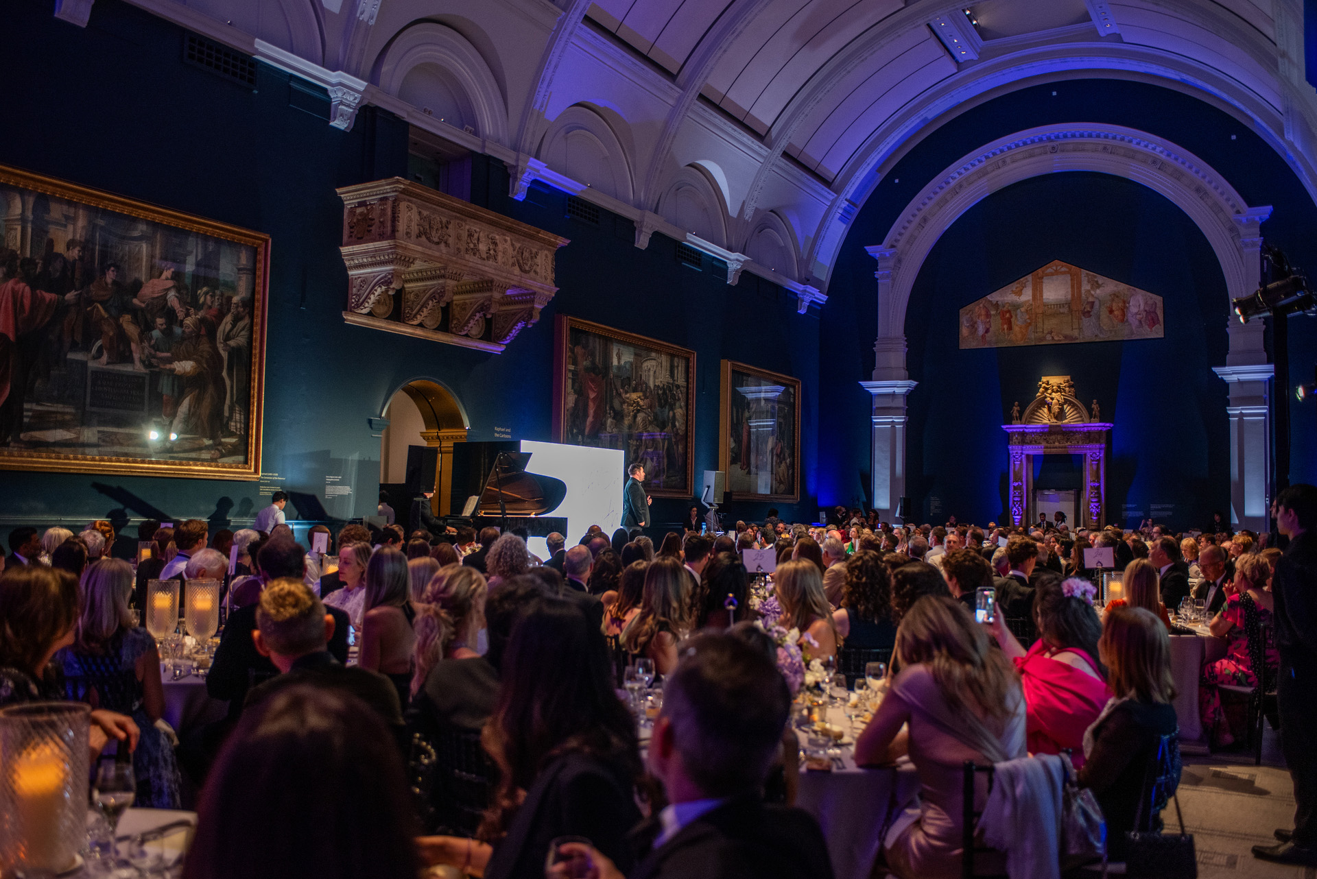 Guests seated at gala dinner tables in a grand gallery hung with Raphael cartoons, watching a speaker on stage beside a piano