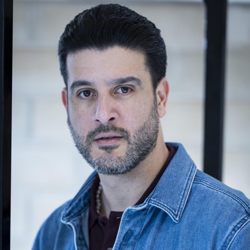 Man with short dark hair and a greying beard, wearing a denim jacket over a maroon top
