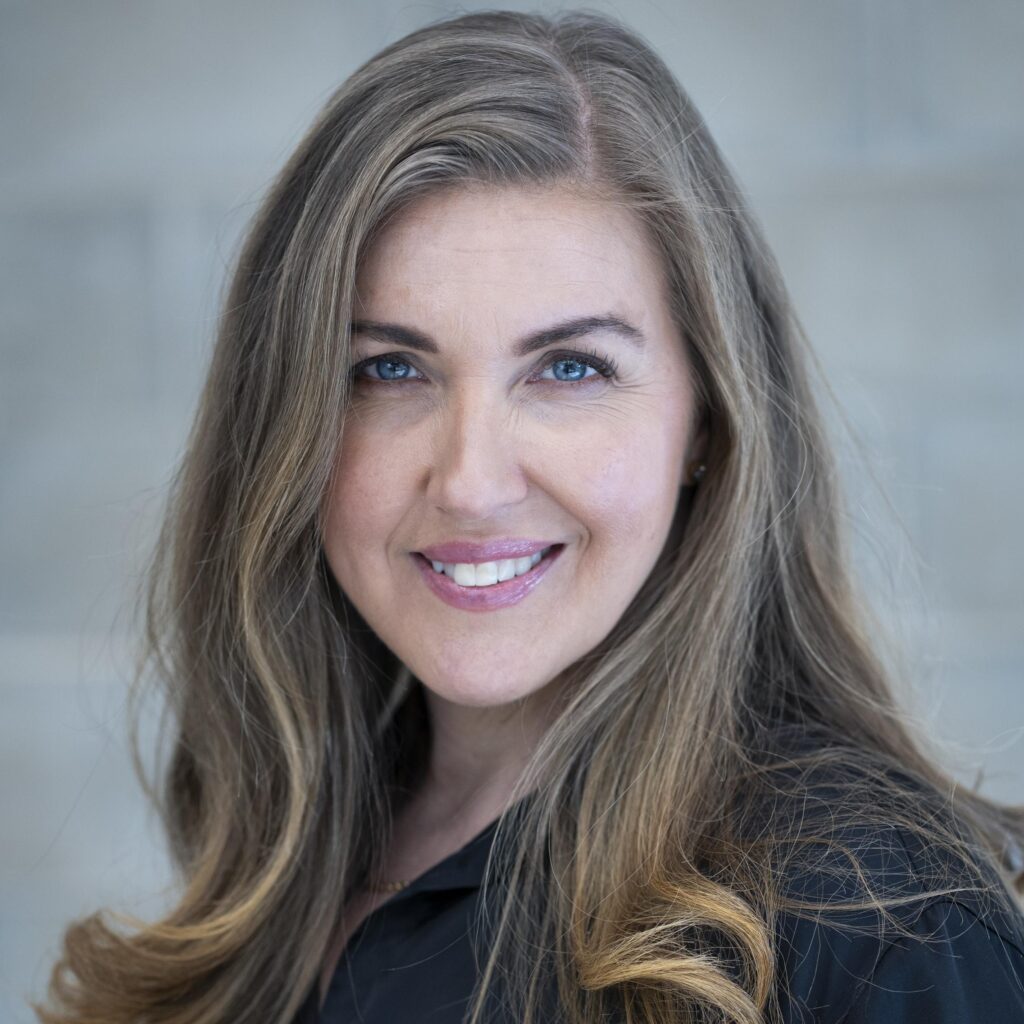 Portrait photo of a woman with long grey and blonde hair, smiling at the camera, wearing a dark top