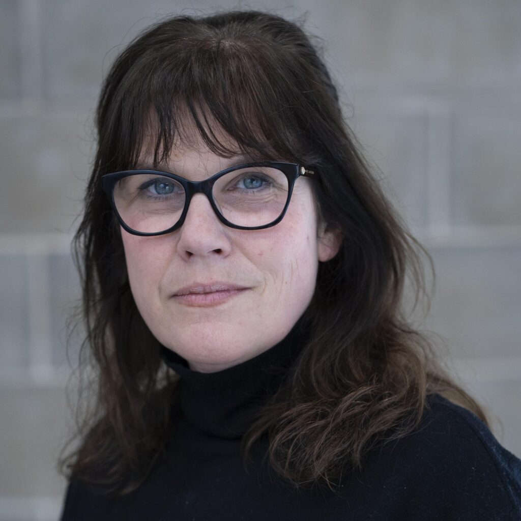 Woman with dark hair and glasses wearing a black polo neck, looking directly at the camera