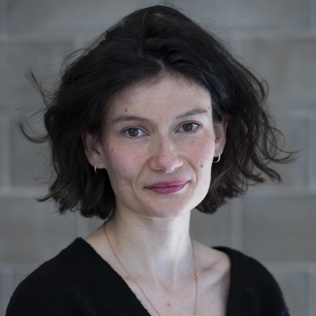 Portrait of a woman with dark wavy shoulder-length hair, wearing a black top against a grey backdrop
