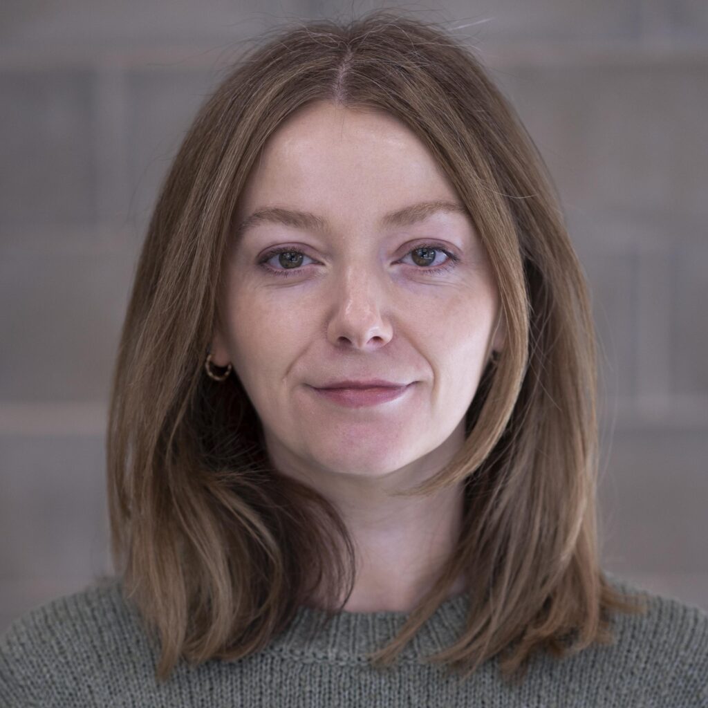 Portrait of a woman with shoulder-length brown hair, wearing a grey jumper, smiling slightly at the camera