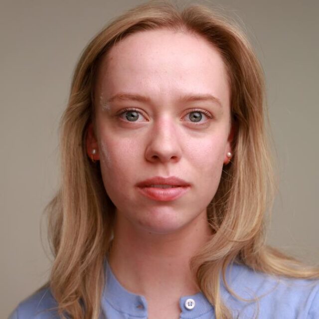 Close-up portrait of a young woman with blonde hair and pearl earrings, wearing a light blue top, looking directly at the camera with a neutral expression